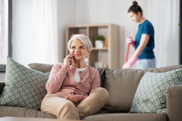 elderly woman sitting on a couch while talking on the phone and a woman behind her cleaning